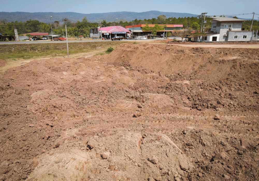 Large expanse of cleared reddish-brown dirt earthwork at a construction site near a rural road with buildings and hills in the background.