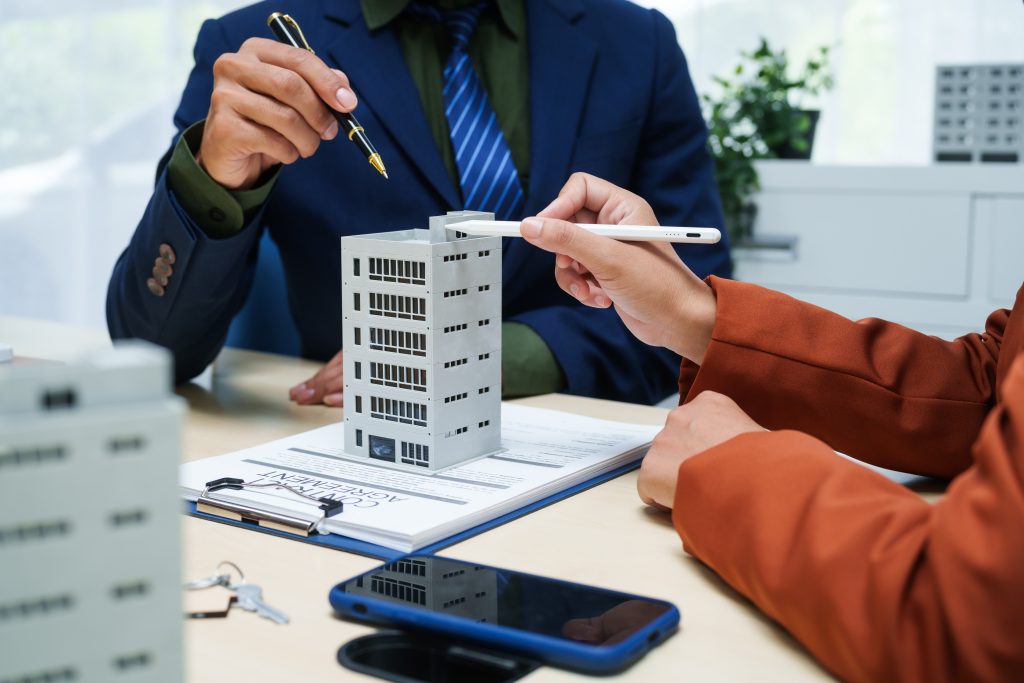 stock image of two people in suits sitting at a desk, using pens to point to a miniature building model that is placed in between them.